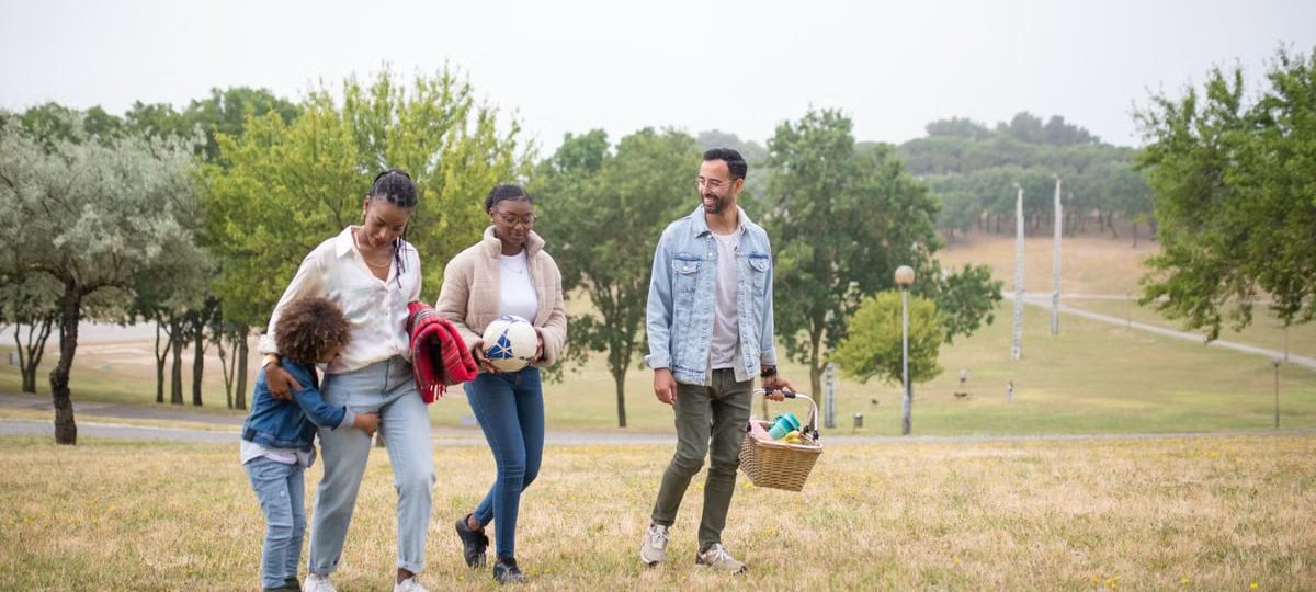 A joyful family walking through a park on a sunny day, carrying a picnic basket and a ball.