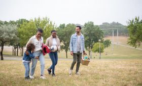 A joyful family walking through a park on a sunny day, carrying a picnic basket and a ball.